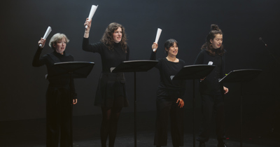 Sur fond noir, vêtues de noir, quatre femmes récitent un texte avec lutrins devant elle, brandissant un texte roulé